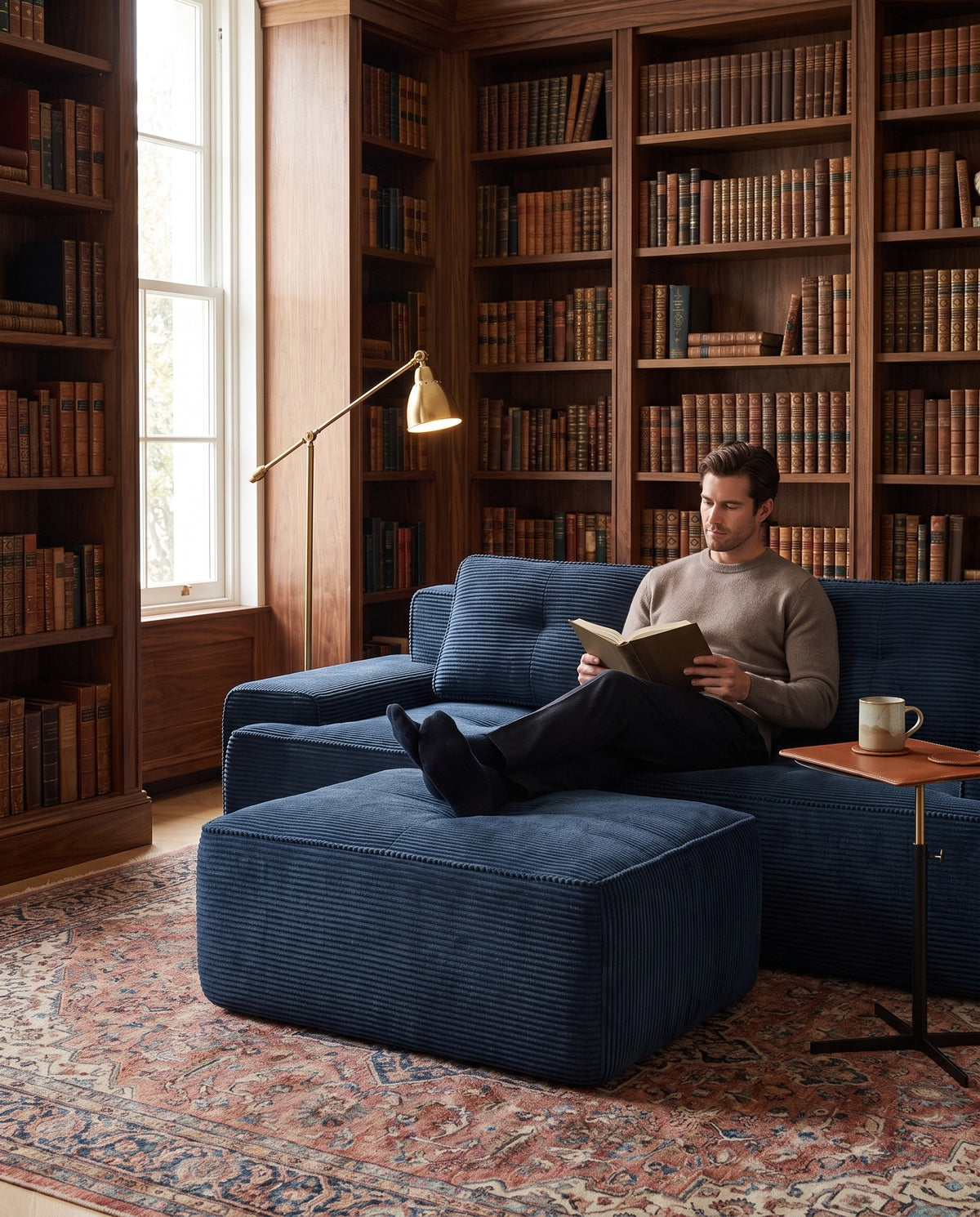 A man reads with his feet up on a navy corduroy modular sofa and ottoman in a library